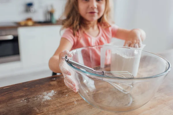 Selective focus of kid mixing flour in measuring jug near whisk in glass bowl while cooking on kitchen table — Stock Photo