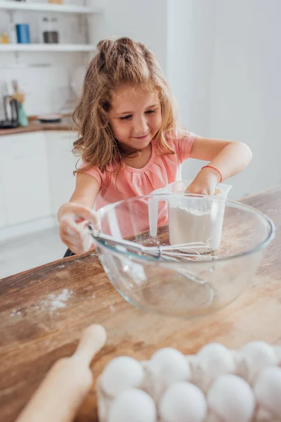 Selective focus of child mixing flour in measuring jug near glass bowl, rolling pin and chicken eggs — Stock Photo