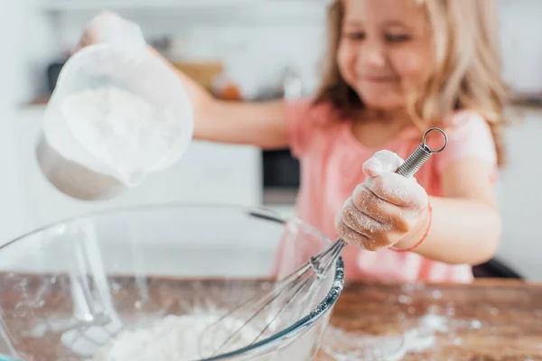 Foco seletivo da menina derramando farinha em tigela de vidro enquanto segurando whisk — Fotografia de Stock