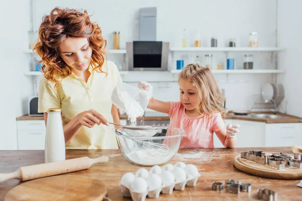 Foco seletivo da jovem mulher encaracolada com a filha peneirando farinha em tigela de vidro perto de ingredientes e cortadores de biscoitos na mesa — Fotografia de Stock