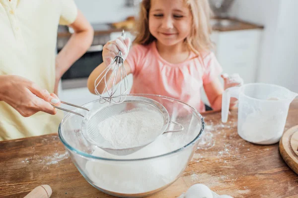 Vista recortada de la madre con tamiz e hija con batidor tamizado harina en tazón de vidrio, enfoque selectivo — Stock Photo