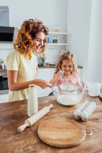 Enfoque selectivo de la madre y la hija tamizar la harina en un recipiente de vidrio cerca de los huevos, la leche, la tabla de cortar y el rodillo - foto de stock