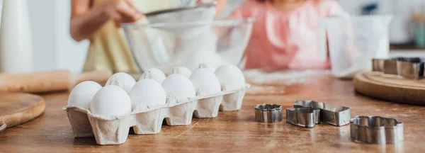 Foco seletivo de ovos de galinha e cortadores de biscoitos perto de mulher e criança peneirando farinha, conceito horizontal — Fotografia de Stock