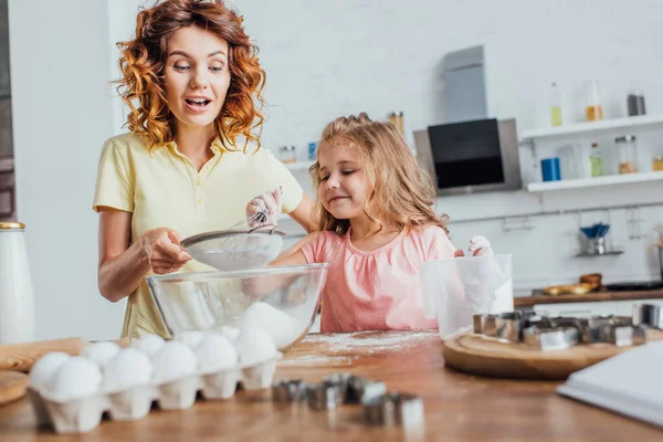 Foyer sélectif de femme bouclée tamiser la farine dans un bol en verre près de la fille, oeufs de poulet et coupe-biscuits — Photo de stock