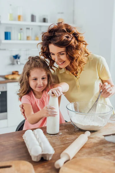 Mulher segurando batedor perto tigela com flor e tocando filha abertura garrafa de leite — Fotografia de Stock