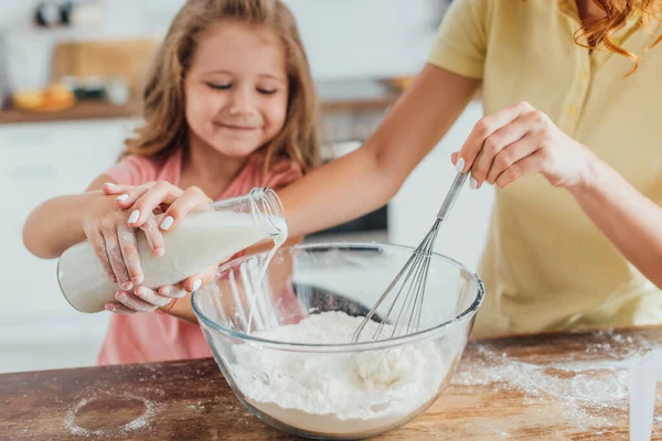 Cropped view of woman kneading dough while pouring milk into glass bowl together with daughter — Stock Photo