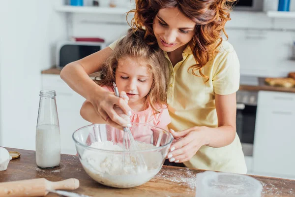 Selective focus of mom kneading dough in glass bow near daughter and bottle of milk — Stock Photo
