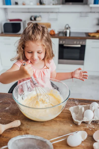 Высокий угол зрения маленькой девочки месить тесто с венчиком в стеклянной миске на кухонном столе — стоковое фото