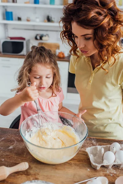 Vista ad alto angolo della ragazza impastare pasta con frusta in ciotola di vetro vicino alla madre — Foto stock