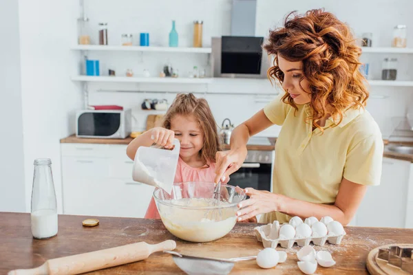 Figlia che aggiunge farina nella ciotola di vetro mentre la madre impasta la pasta — Foto stock