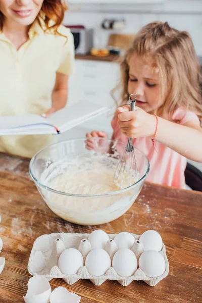 Vista ritagliata della madre che legge il ricettario mentre la figlia impasta la pasta con la frusta nella ciotola di vetro, messa a fuoco selettiva — Foto stock