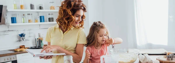 Horizontales Konzept: Mutter hält Kochbuch in der Hand, während Tochter Teig in Glasschüssel zubereitet — Stockfoto