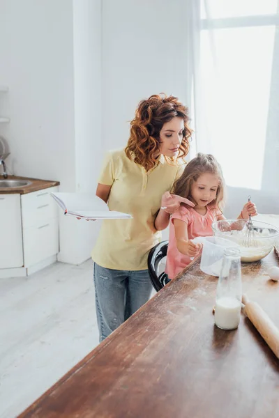 Enfoque selectivo de la madre sosteniendo libro de cocina y señalando con el dedo cerca de la hija amasando masa en un tazón de vidrio - foto de stock