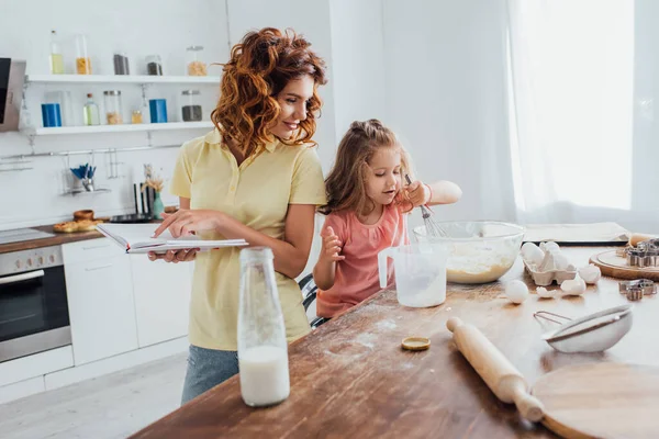 Selektiver Fokus der Mutter, die mit dem Finger auf Kochbuch zeigt, während Tochter Teig in Glasschüssel knetet — Stockfoto