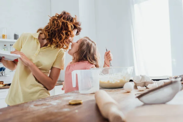 Enfoque selectivo de la madre sosteniendo platos mientras está de pie cara a cara con la hija cerca de la mesa con utensilios de cocina - foto de stock