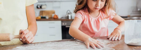Vue recadrée de la mère debout près de sa fille dispersant de la farine sur une table en bois dans la cuisine — Photo de stock