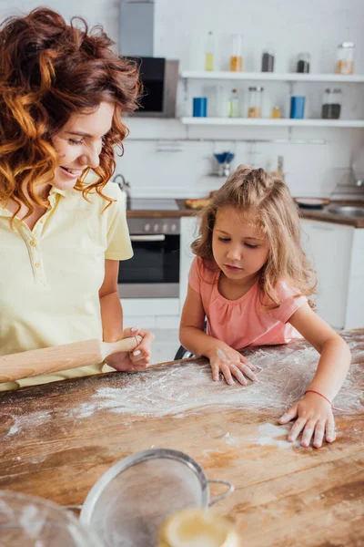 Foco seletivo da menina espalhando farinha na mesa da cozinha perto da mãe segurando rolo pin — Fotografia de Stock