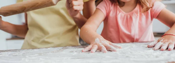 Vista parcial de la hija dispersando harina en la mesa de la cocina cerca de la madre sosteniendo el rodillo, tiro panorámico - foto de stock