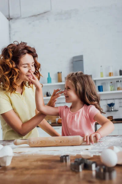 Вибірковий фокус матері і дочки торкаються носів один одного біля прокатного штифта і тіста на кухонному столі — стокове фото