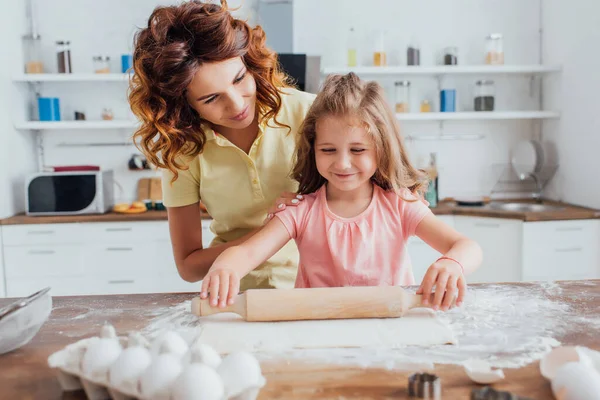 Enfoque selectivo de la madre tocando hombros de hija desplegando la masa en la mesa dispersa con harina - foto de stock