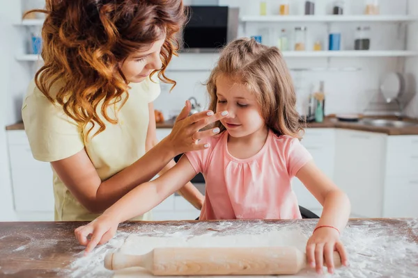 Mother touching nose of daughter rolling out dough on table scattered with flour — Stock Photo