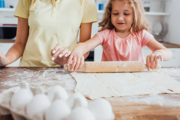 Vista ritagliata della madre vicino figlia stendere pasta sul tavolo sparsi con farina — Foto stock