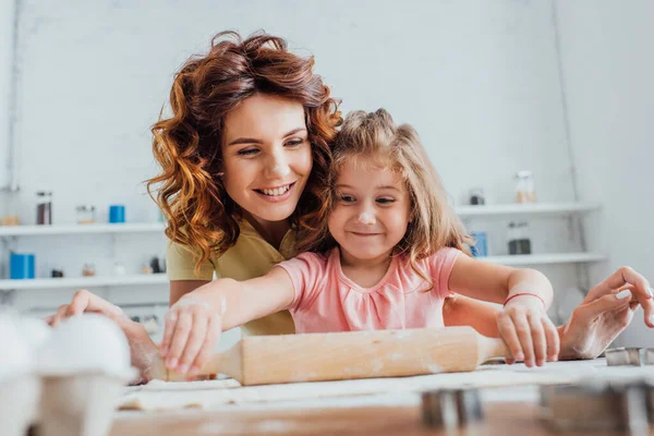 Foyer sélectif de l'enfant déroulant la pâte sur la table près de la jeune mère bouclée — Photo de stock