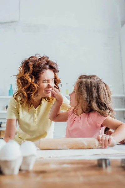 Selective focus of daughter touching nose of mother while rolling out dough on table — Stock Photo