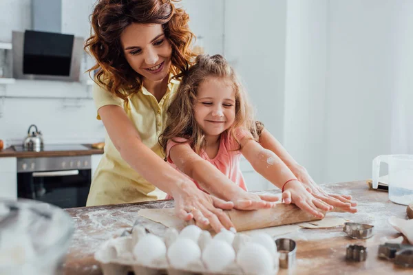 Enfoque selectivo de la madre y su hija el despliegue de masa cerca de huevos de pollo y cortadores de galletas en la mesa - foto de stock