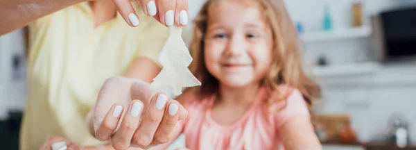 Vista parcial de la mujer sosteniendo la galleta cruda en forma de árbol cerca de hija y papel de hornear, imagen horizontal - foto de stock