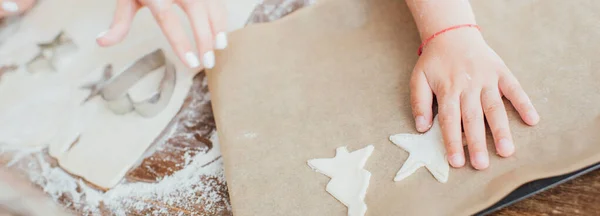 Vue partielle de l'enfant mettant des biscuits multiformes crus sur du papier cuisson près de la mère et de la pâte roulée, prise de vue panoramique — Photo de stock