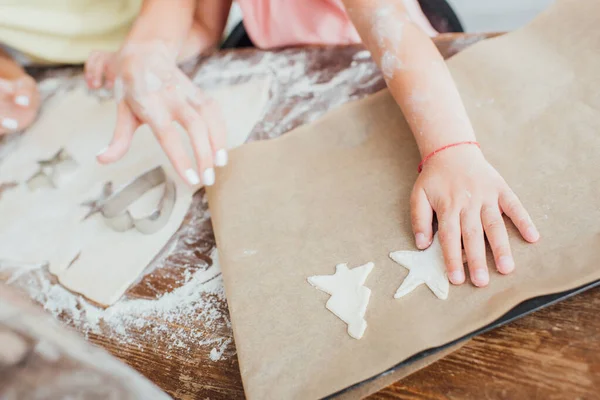 Vista recortada del niño poniendo galletas crudas multiformes en papel de hornear cerca de la madre y la masa enrollada, enfoque selectivo - foto de stock