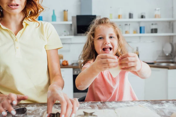 Selective focus of blonde girl holding raw cookie while mother cutting rolled dough with molds — Stock Photo