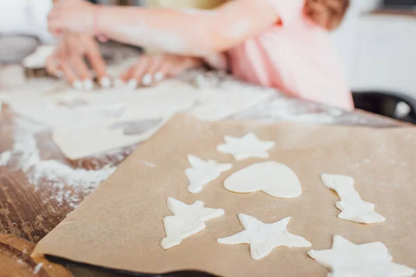 Vista recortada de la madre y la hija cerca de papel de hornear con galletas crudas en forma de múltiples en la mesa de la cocina, enfoque selectivo - foto de stock