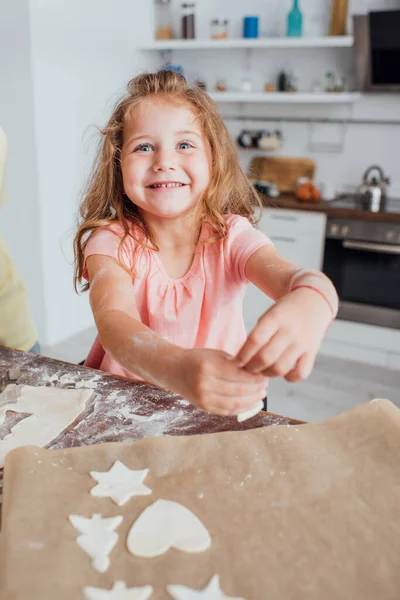 Enfoque selectivo de galletas crudas en múltiples formas sobre papel de hornear cerca de niña rubia - foto de stock