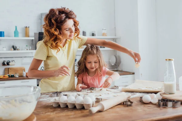 Enfoque selectivo de la joven madre y su hija preparando galletas cerca de huevos de pollo, leche y utensilios de cocina en la mesa de la cocina - foto de stock