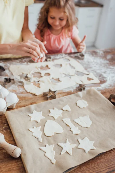 Enfoque selectivo de galletas crudas en múltiples formas en papel de hornear cerca de mamá y su hija - foto de stock