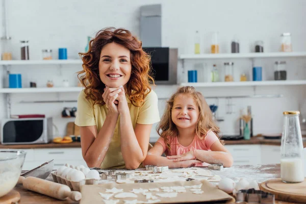 Junge Mutter und Tochter schauen in die Kamera neben rohen Keksen und Zutaten auf dem Küchentisch — Stockfoto