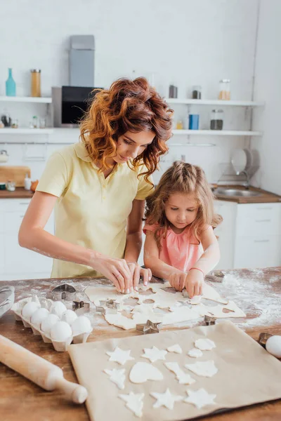 Selective focus of curly mother and blonde daughter cutting out cookies from rolled dough — Stock Photo
