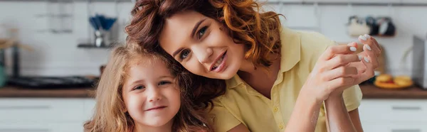 Horizontal image of curly mother and blonde daughter looking at camera in kitchen — Stock Photo