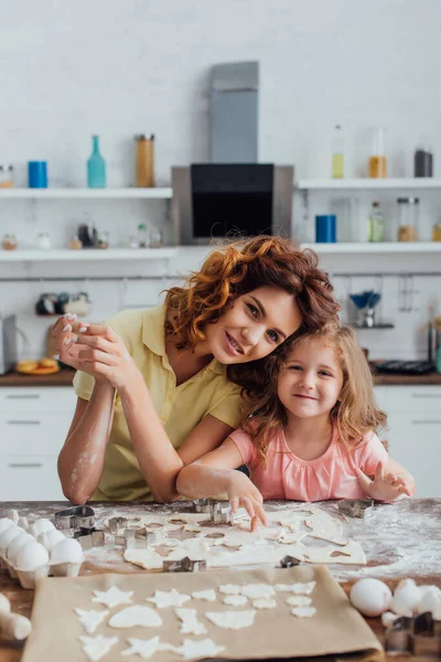 Foyer sélectif de la fille blonde et la mère bouclée regardant la caméra près de la pâte et des biscuits multi-formes sur papier cuisson — Photo de stock