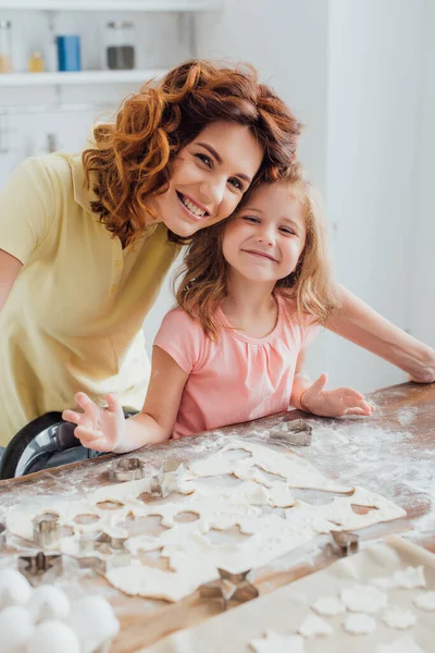 Selektiver Fokus der lockigen Mutter und der blonden Tochter, die in die Kamera neben Teig schauen und Kekse schneiden — Stockfoto