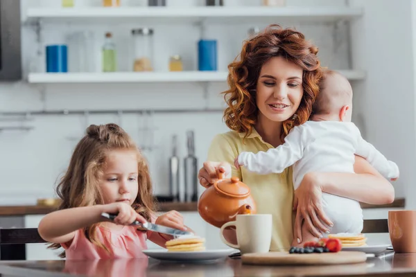 Selective focus of mother pouring tea from teapot while holding infant near daughter eating pancakes — Stock Photo