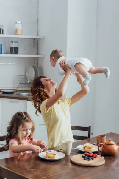 Giovane madre tenendo bambino ragazzo sopra la testa vicino figlia mangiare frittelle per la prima colazione — Foto stock