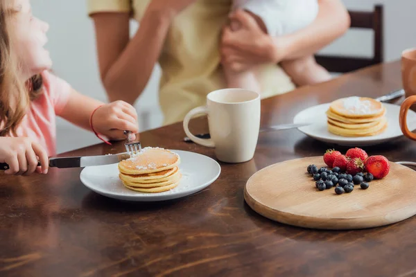 Vista recortada de la madre sosteniendo al hijo pequeño cerca de la hija comiendo deliciosos panqueques, enfoque selectivo - foto de stock