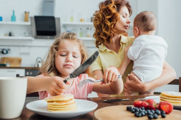 Messa a fuoco selettiva della ragazza scontenta con forchetta e coltello vicino ai pancake mentre la madre tiene in braccio il bambino — Foto stock