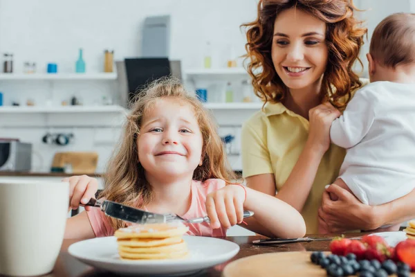 Selective focus of girl looking at camera while eating pancakes near mother with infant — Stock Photo