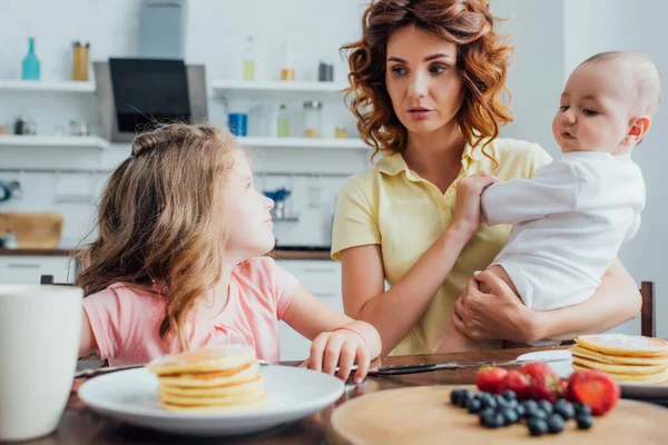 Selektiver Fokus der Mutter, die ihren Sohn hält, während sie die Tochter neben dem Teller mit leckeren Pfannkuchen sitzen sieht — Stockfoto
