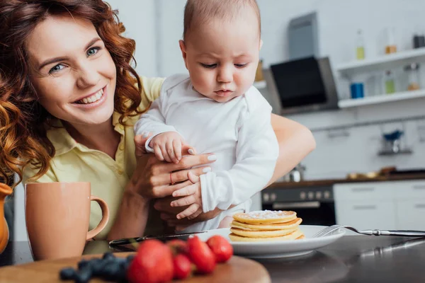 Messa a fuoco selettiva della giovane madre guardando la fotocamera mentre tiene in braccio il figlio neonato vicino a deliziosi pancake — Foto stock