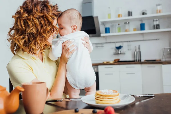 Foyer sélectif de jeune mère bouclée tenant son fils près de la table servi avec de délicieuses crêpes — Photo de stock